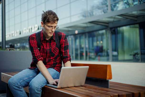 Ένας αφοσιωμένος έξυπνος φοιτητής κάθεται σε ένα παγκάκι στην πανεπιστημιούπολη και πληκτρολογεί σε ένα λάπτοπ. Πορτρέτο ενός φοιτητή πανεπιστημίου που κάθεται σε ένα παγκάκι και εργάζεται για τη σχολική του εργασία εξ αποστάσεως. - Φωτογραφία, εικόνα