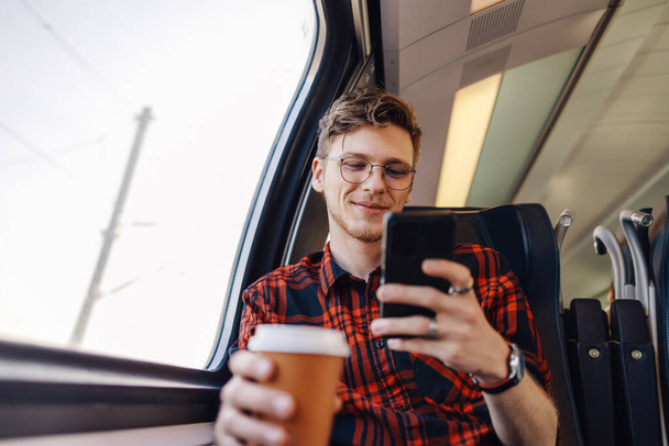 Nuori matkustaja istuu junassa ikkunan vieressä ja kirjoittaa viestejä puhelimeen hymyillen sille. Työmatkalainen lähtee matkalle junalla ja kirjoittaa puhelimeen kahvin äärellä.. - Valokuva, kuva