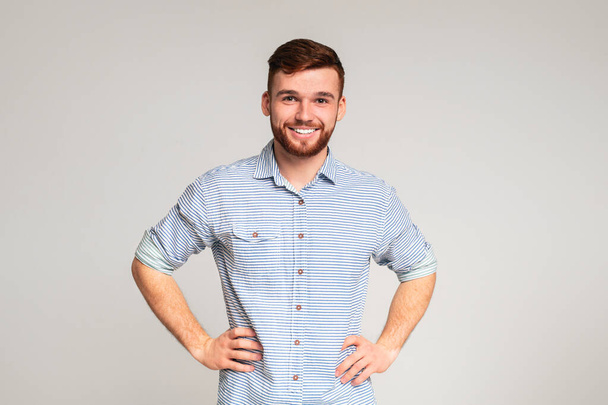 Портрет улыбающегося парня в повседневной рубашке с руками на бёдрах на сером фоне студии, копировальное пространство - Фото, изображение