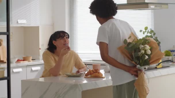 Mediano largo tiro de hombre afroamericano dando flores a su esposa de etnia caucásica durante el desayuno en casa - Imágenes, Vídeo
