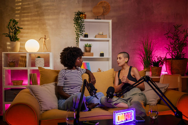 Afrykanka treści twórca wywiady słynnej białej kobiety w przytulnym studio siedząc na żółtej kanapie. Nagrywanie podcastu internetowego przy użyciu profesjonalnego mikrofonu i sprzętu audio. - Zdjęcie, obraz