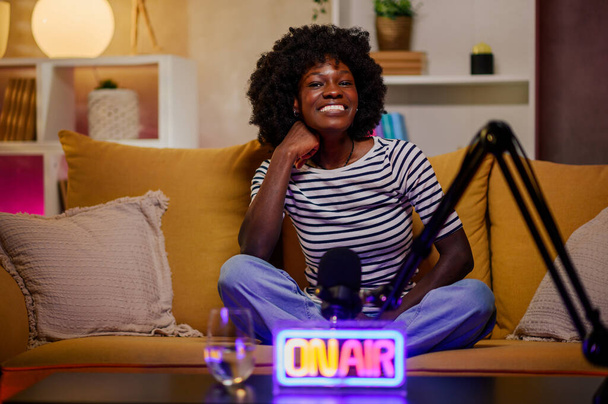 Young african american woman smiling while recording an audio broadcast in a modern cozy studio. Cheerful black female content creator hosting an internet podcast. Copy space. Looking into the camera. - Photo, Image