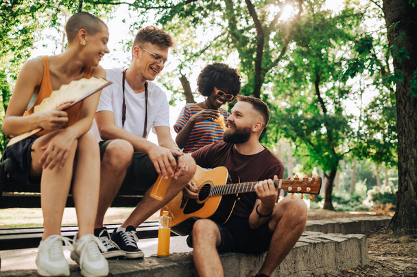 Çok kültürlü sokak sanatçılarından oluşan küçük bir grup parkta oturup reçel seansı yapıyorlar. Diğerleri dinlerken bir adam gitar çalıyor. Doğada arkadaşlarıyla prova yapan bir adam.. - Fotoğraf, Görsel