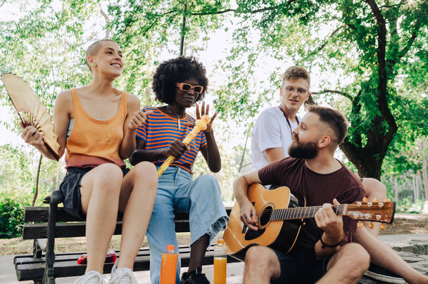 Группа мультикультурных уличных артистов, сидящих в парке в солнечный летний день и репетирующих. Мужчина играет на гитаре, в то время как африканская американка поет песню. Путешествующие художники в парке. - Фото, изображение