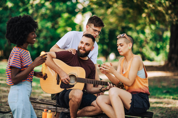 Vier multirassische Influencer versammelten sich im Park, um gemeinsam abzuhängen und Inhalte füreinander zu schaffen. Vier Freunde spielen Gitarre, haben Spaß und fotografieren an sonnigen Tagen draußen. Kopierraum. - Foto, Bild