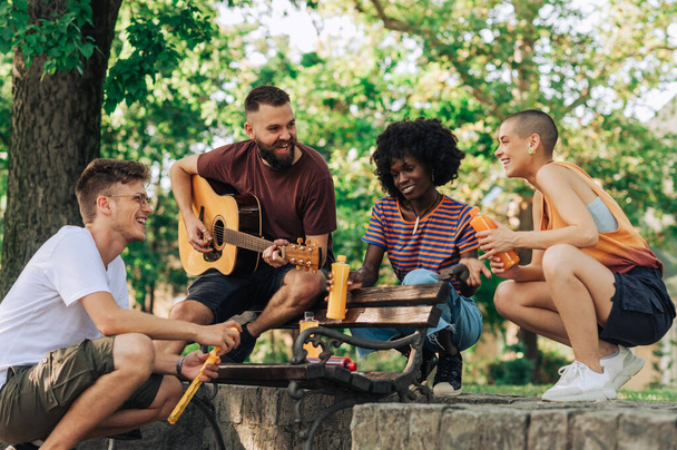 Группа из четырех молодых людей, сидящих на скамейке и поющих под гитарную музыку и пьющих безалкогольные напитки. Молодые люди играют на гитаре в парке и тусуются вместе. - Фото, изображение