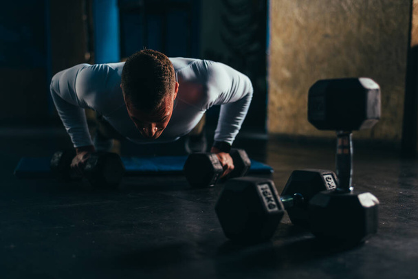 Muscular handsome man doing pushups with dumbbells while training in the gym - Photo, Image
