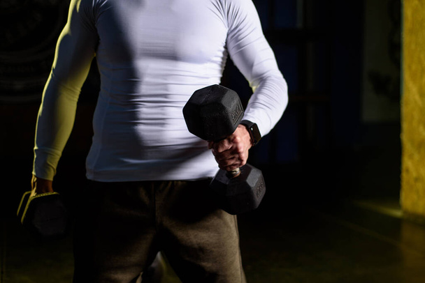 Молодой привлекательный мужчина держит гантели за руки во время тренировки в спортзале - Фото, изображение