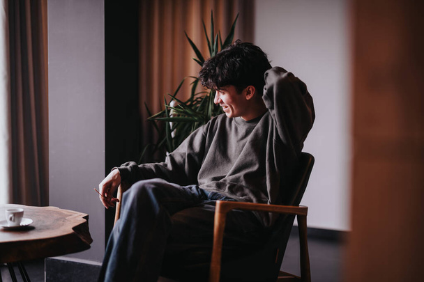 Eine gemütliche Szene, in der ein junger Mann lächelt und es sich zu Hause auf einem Stuhl gemütlich macht, mit einer warmen und einladenden Atmosphäre. - Foto, Bild