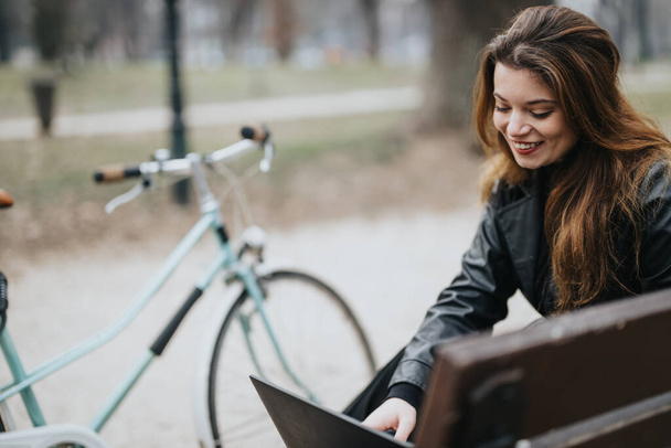 Mujer joven elegante y segura de sí misma utilizando un ordenador portátil al aire libre, entorno de negocios casual con bicicleta, que encarna un estilo de vida moderno y activo. - Foto, imagen