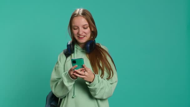 Portret szczęśliwej nastolatki czytającej wiadomości przez telefon, zachwycony po otrzymaniu dobrych wiadomości. Radosna kobieta podekscytowana SMS na smartfonie, świętowanie, tło studio, aparat A - Materiał filmowy, wideo