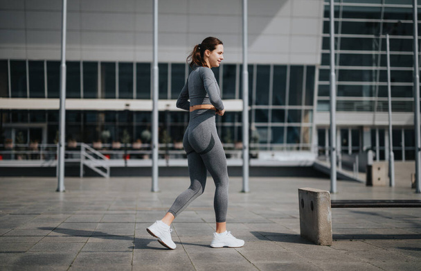 Πλευρική άποψη μιας νεαρής γυναίκας με αθλητικά ρούχα που τρέχει σε ένα περιβάλλον πόλης, επιδεικνύοντας έναν υγιεινό τρόπο ζωής - Φωτογραφία, εικόνα