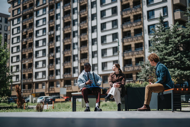 Three young professionals engage in a casual business discussion outdoors, seated on benches with a modern city apartment building in the background. - Photo, Image
