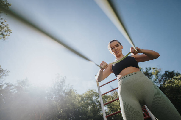 Fit woman performing resistance band exercises outside in park. Concept of outdoor fitness, strength training, and healthy lifestyle. - Photo, Image