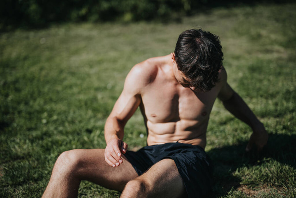 Muskelkräftiger, hemdsloser Mann, der nach einem anstrengenden Training in der Sonne im Gras in einem Park sitzt und sich ausruht. Fitness und gesunder Lebensstil. - Foto, Bild