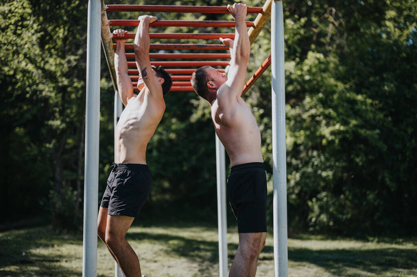 Dwóch młodych mężczyzn ćwiczy na barach dla małp w plenerze parku, ciesząc się słoneczną pogodą i aktywnością fizyczną. - Zdjęcie, obraz