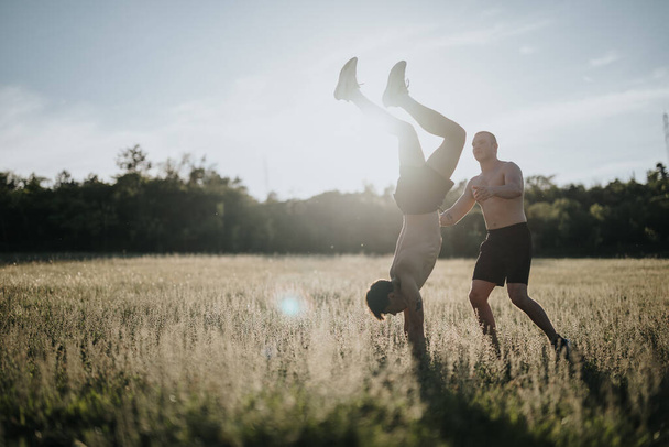 Двое мужчин занимались спортом, тренировались на руках на безмятежном солнечном поле. Изображение изображает движение, фитнес и природную красоту. - Фото, изображение