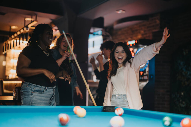 Joyful friends gather around a billiard table for a night out at a bar, displaying happiness and togetherness. A woman celebrates a successful shot. - Photo, Image