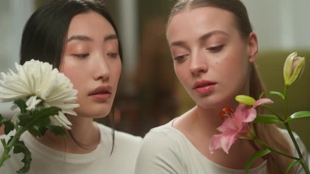 Portré két barát nők arc szépség vonzó egészséges női lányok nők multiracionális többnemzetiségű kaukázusi nő és ázsiai lány japán kínai koreai virágokkal természet kozmetikai szerves - Felvétel, videó