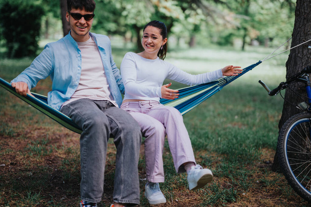 Ένα χαρούμενο νεαρό ζευγάρι χαλαρώνει σε μια μπλε αιώρα, γελώντας μαζί σε ένα καταπράσινο αστικό πάρκο σε μια όμορφη ηλιόλουστη μέρα. - Φωτογραφία, εικόνα