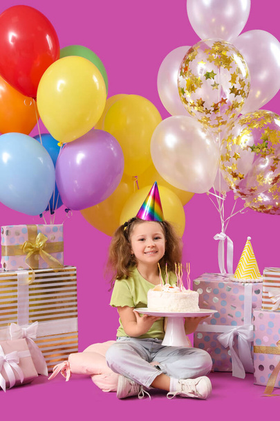 Linda niña en sombrero de fiesta con globos, pastel de cumpleaños y cajas de regalo sobre fondo púrpura - Foto, Imagen