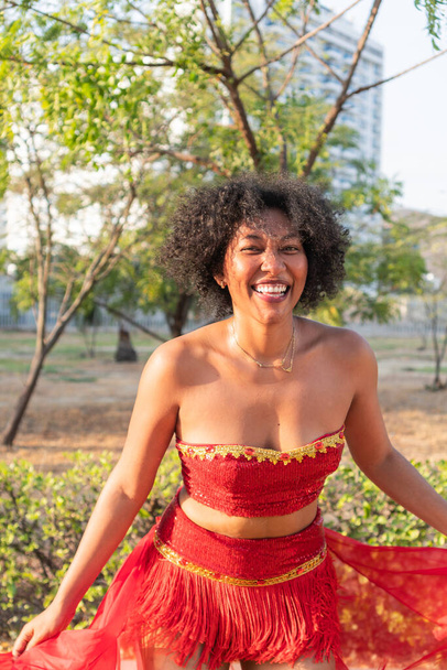 Uśmiechnięta kobieta w czerwonej sukience cieszy się słonecznym dniem w miejskim parku, otoczona zielenią i wieżowcami, uchwycając radosną i beztroską chwilę. - Zdjęcie, obraz