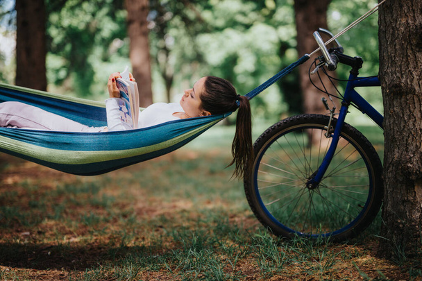 Eine friedliche Szene in einem üppigen Park, wo eine junge Frau in einer Hängematte liegt, tief in ein Buch vertieft, mit ihrem Fahrrad in der Nähe geparkt. - Foto, Bild