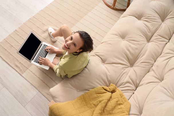 Mujer joven con portátil sentado en el suelo cerca del sofá en la sala de estar - Foto, imagen