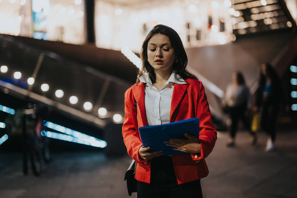 Joven mujer profesional en una chaqueta roja revisando documentos al aire libre por la noche con luces de la ciudad en el fondo. - Foto, imagen