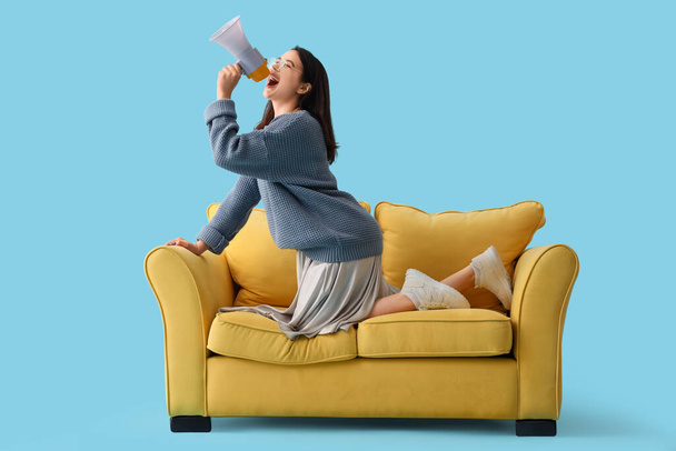 Junge Frau brüllt in Megafon auf gelbem Sofa vor blauem Hintergrund - Foto, Bild
