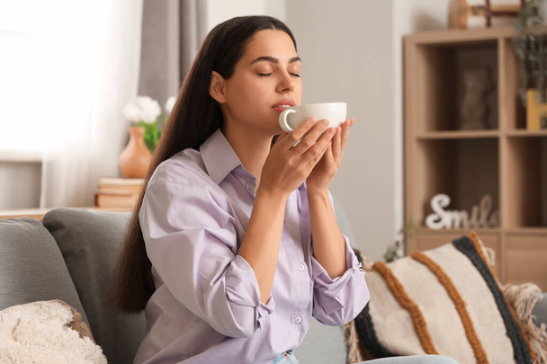 Csinos fiatal nő csésze forró kávé ül kanapén otthon - Fotó, kép