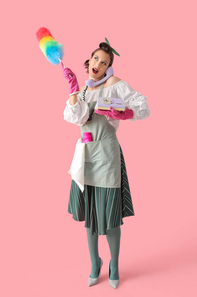 Молодая шокированная домохозяйка с пылесосом и моющим средством разговаривает по телефону на розовом фоне - Фото, изображение
