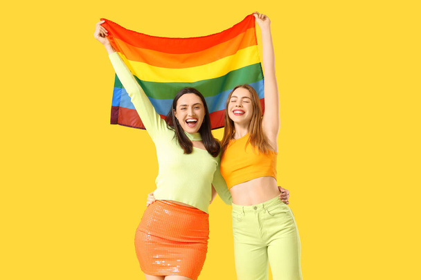 黄色の背景にLGBTの旗を持つ美しい若いレズビアンカップル - 写真・画像