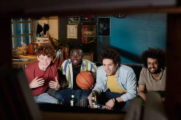 Skupina různých mužských přátel sedících na gauči a sledujících basketbalový zápas v televizi pozdě večer - Fotografie, Obrázek