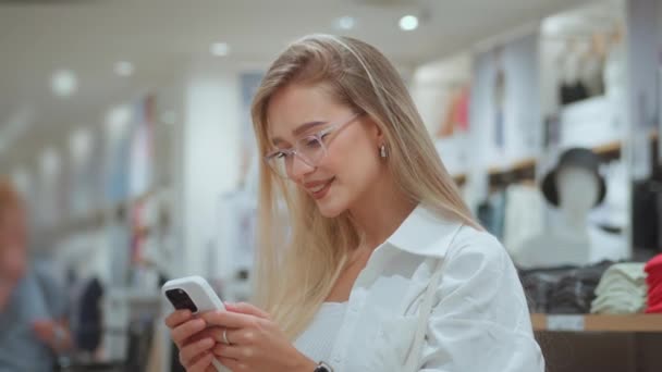 Közepes fehér nő áll ruhaboltban a bevásárlóközpontban mosolyogva a mobiltelefonján. Millenniumi kaukázusi nő szemüvegben áll ruhaüzlet bevásárló komplexum mosolygós telefon - Felvétel, videó
