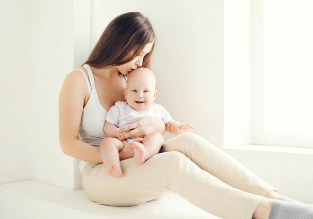 Lovely young mom kissing her baby at home in light room - Zdjęcie, obraz