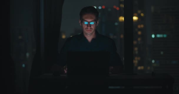 Home office mid adult person in glasses sitting desk works on laptop late night front view. Caucasian millennial man eyeglasses sits table working computer nighttime work dedication focus work - Footage, Video