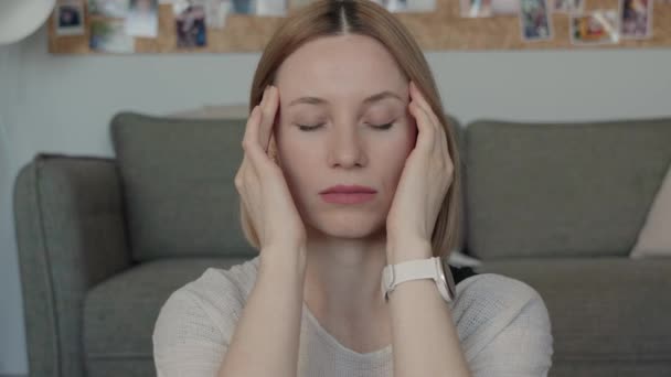 Mujer joven con los ojos cerrados y las manos en las sienes, expresando estrés o pensamiento profundo mientras está sentada en un sofá en casa. - Metraje, vídeo
