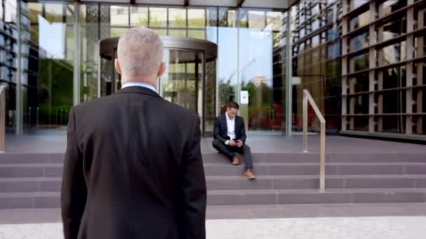 Twee blanke zakencollega 's begroeten elkaar bij de ingang van het kantoorgebouw. Zakenman loopt naar het kantoorgebouw waar hij werkt - Video