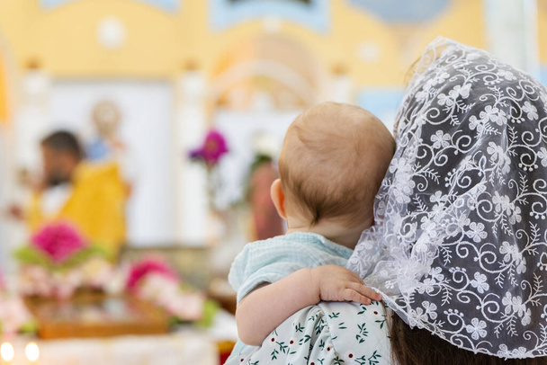 Вид ззаду жінки в білому хустці тримає дитину проти ікон в церкві.  - Фото, зображення