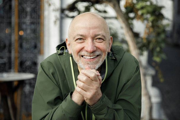 Senior Man met baard vrolijk klampen handen in groene jas buiten - Foto, afbeelding