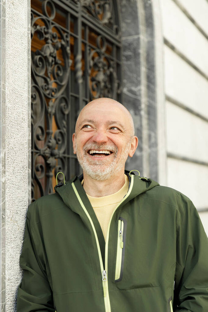 Hombre mayor alegre en chaqueta verde mirando hacia arriba cerca de puerta de metal adornado, concepto de estilo de vida - Foto, imagen