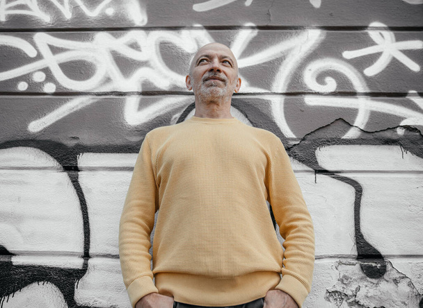Homem sênior com uma barba em uma camisola amarela de pé com as mãos nos bolsos contra uma parede decorada com grafite, olhando para cima - Foto, Imagem
