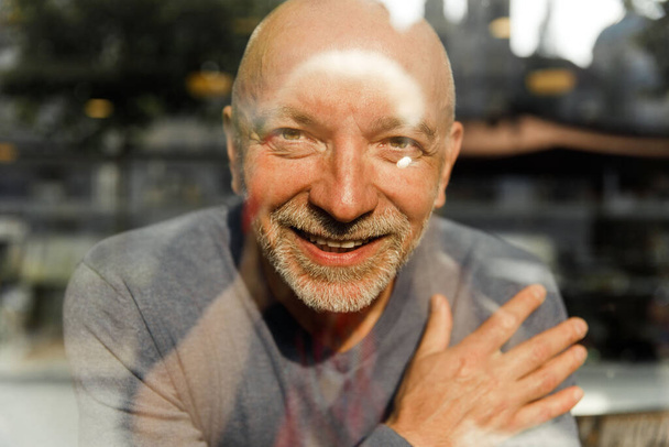 Feliz hombre mayor con barba sonriendo a través de una ventana de cristal, reflejos visibles - Foto, imagen