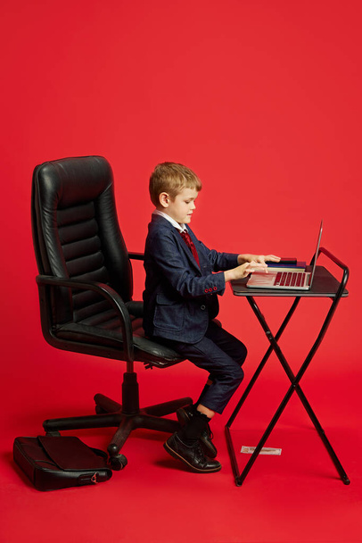 Young boy plays role of businessman against vibrant red studio background. Children dreaming big and exploring different occupations. Concept of children dreams, future career, games, fantasy. Ad - Foto, immagini