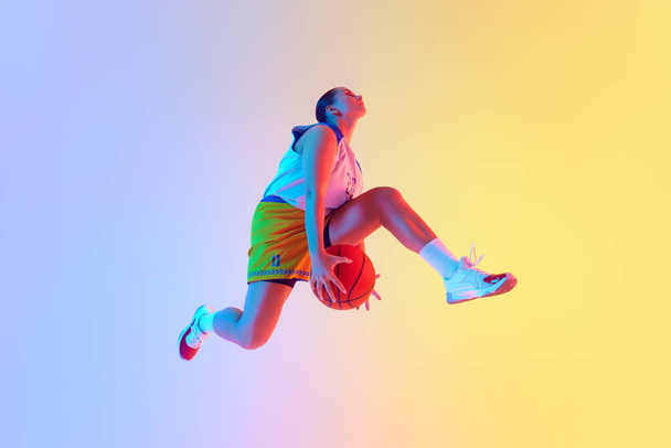 Retrato dinámico de una jugadora de baloncesto que realiza un salto perfecto en el aire para marcar gol en luz de neón contra el fondo del estudio de gradiente. Concepto de deporte, campeonato, torneo, hobby. Anuncio - Foto, Imagen
