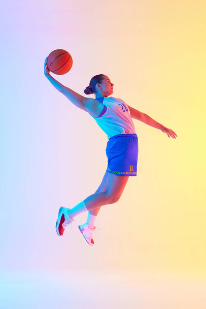 Portret profesjonalnej koszykarki skoki kobiet z piłką gotowy strzelić gola w neonowym świetle na tle gradientowego studia. Pojęcie sportu, mistrzostwa, turniej, hobby, rekreacja - Zdjęcie, obraz