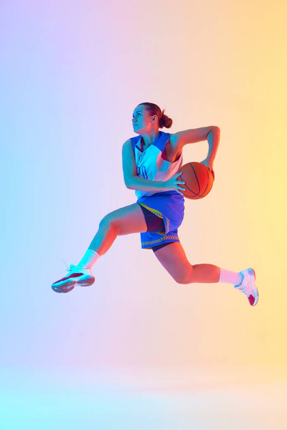 Żeński koszykarz w niebieskiej koszulce przygotowuje się do perfekcyjnego skoku w powietrzu w neonowym świetle na tle gradientowego studia. Pojęcie sportu, turnieju, hobby i rekreacji. Ogłoszenie - Zdjęcie, obraz