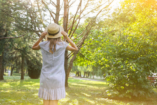 Κομψό χίπστερ κορίτσι με ψάθινο καπέλο στέκεται έξω. Ευτυχισμένη γυναίκα με μπλε φόρεμα. Ταξιδιωτική ιδέα διακοπών. Φωτογραφία καλοκαιρινού τρόπου ζωής. Αποχρώσεις ήλιου και δέντρων. - Φωτογραφία, εικόνα