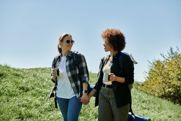 Twee jonge vrouwen, een lesbisch stel, wandelen samen op een zonnige dag. Ze glimlachen en houden elkaars hand vast, genieten van hun tijd in de wildernis.. - Foto, afbeelding
